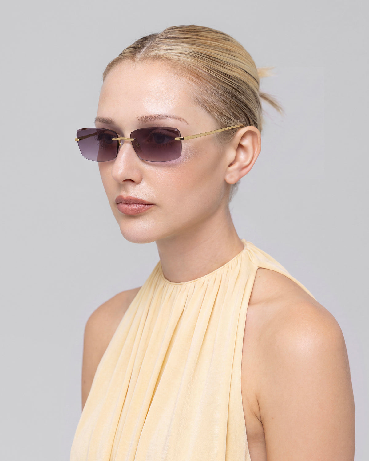 Blonde woman with low bun wears For Art’s Sake® Sol I sunglasses and a sleeveless light beige top, posing against a plain light gray background.