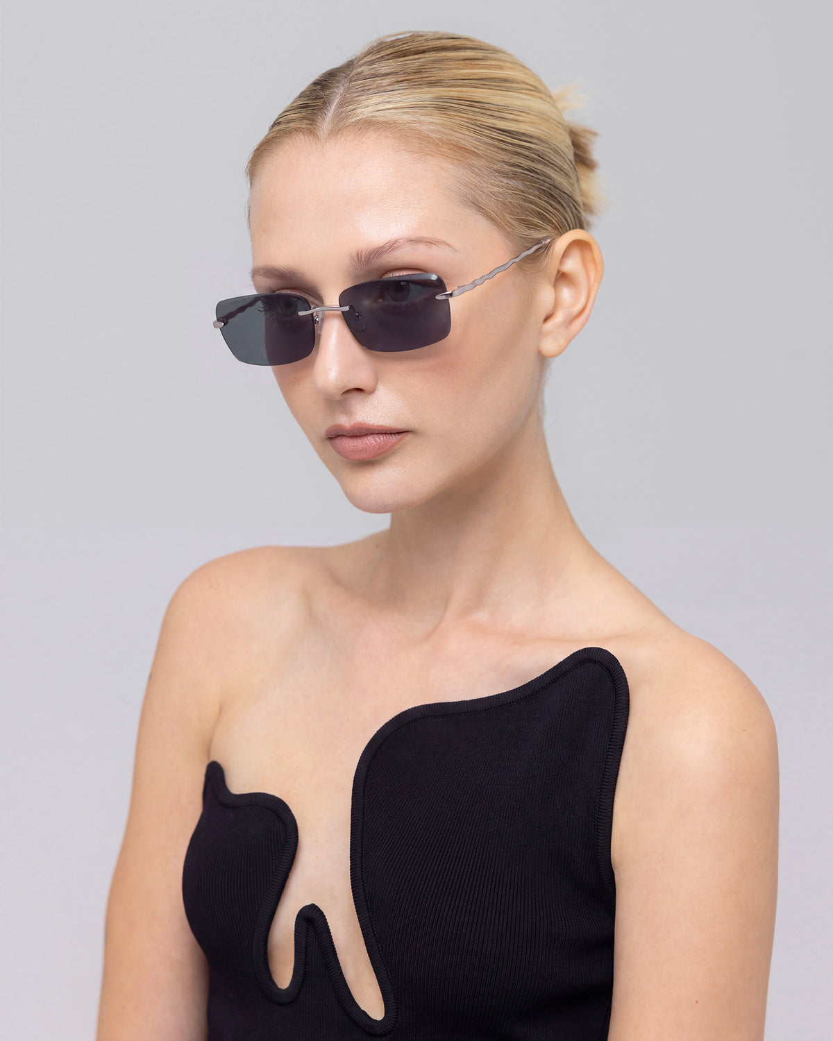 A woman with blonde hair in a low bun wears For Art’s Sake® Sol I sunglasses and a black asymmetrical top, posing against a plain gray background.