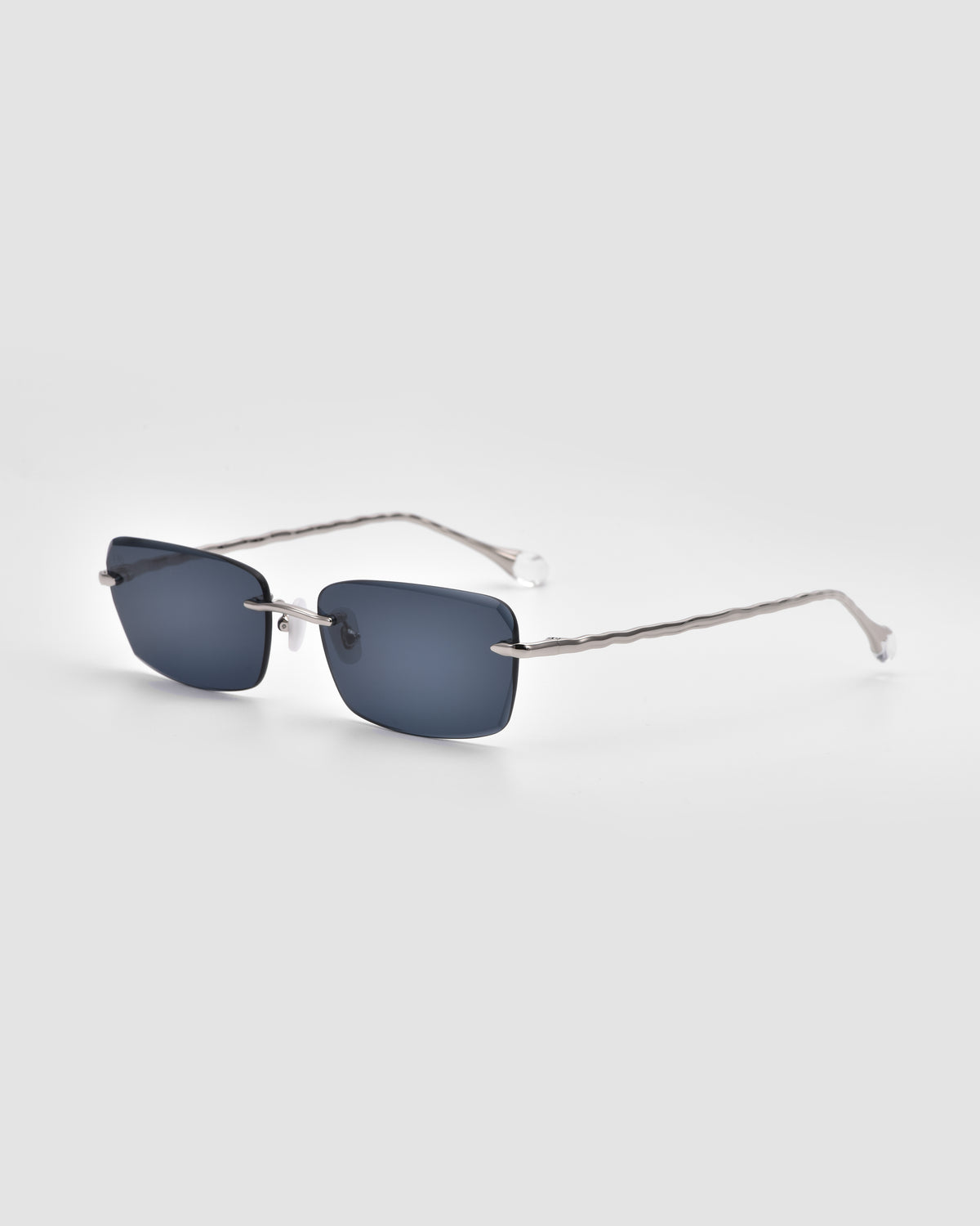 Sol I square sunglasses from For Art’s Sake® feature rimless dark blue lenses, sleek silver metal arms, and sculpted nylon temple tips, displayed on a light gray background.