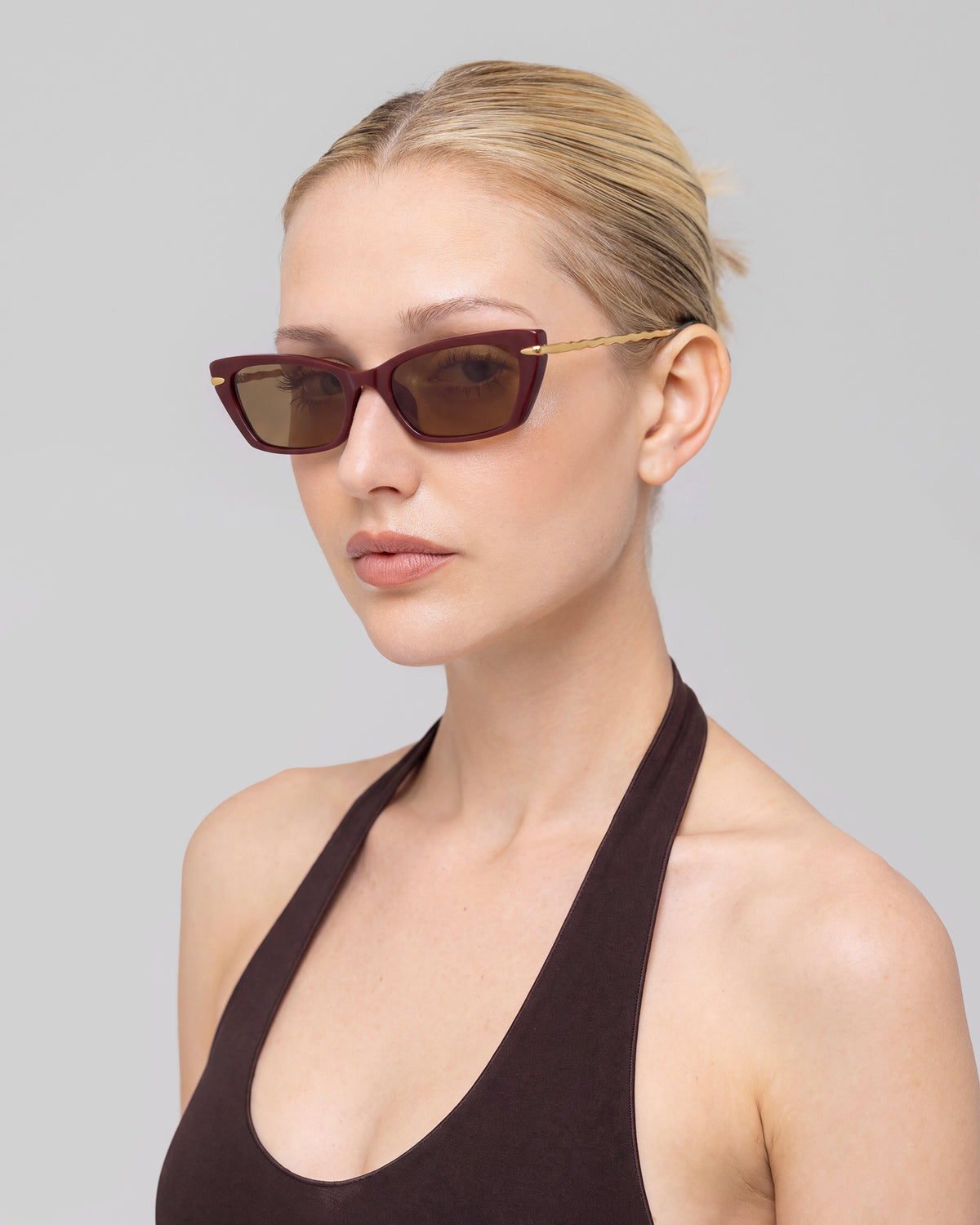 A woman with blonde hair in a low bun wears For Art’s Sake® Sol II sunglasses and a sleeveless dark halter top, standing against a plain light gray background.