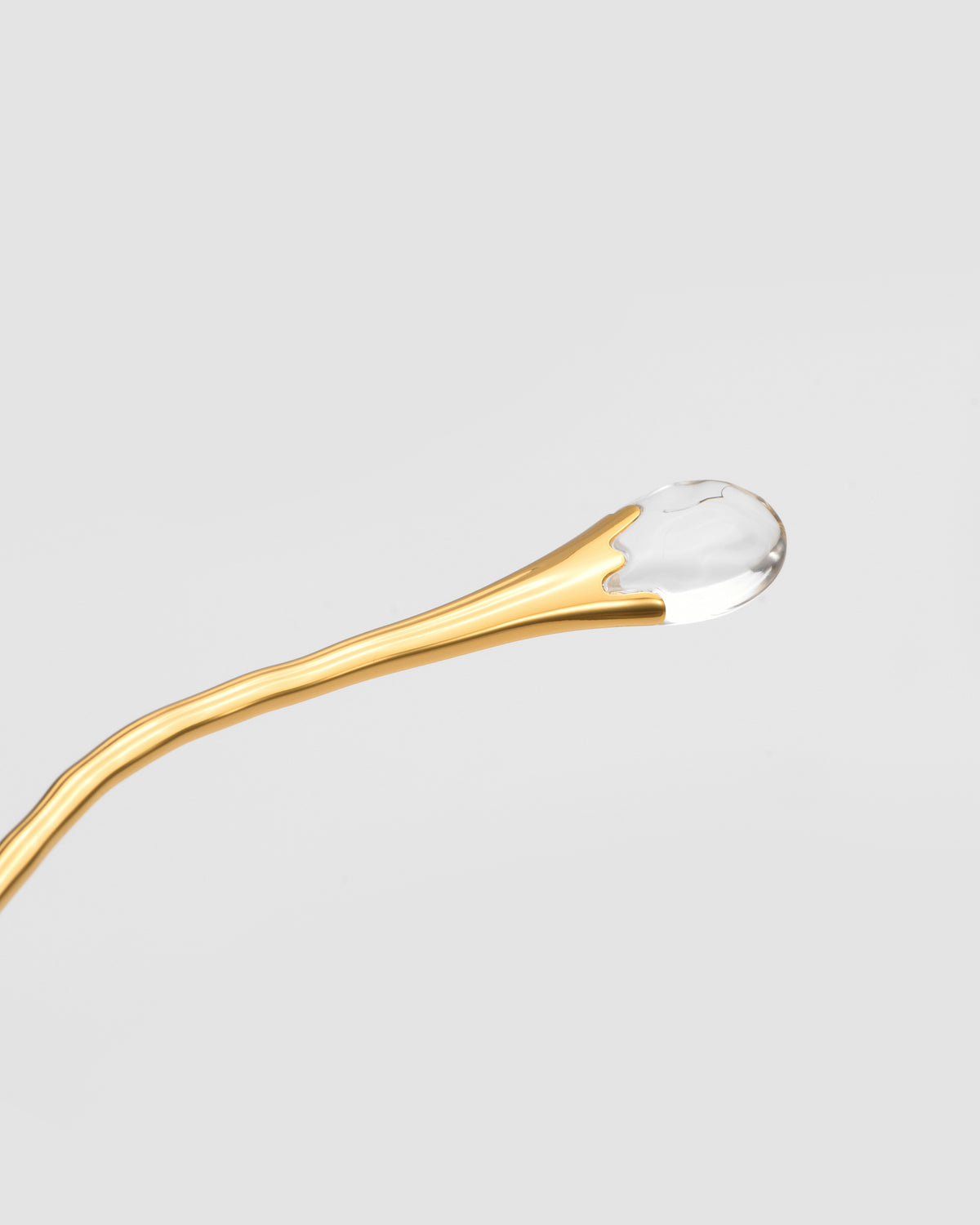 A golden spoon with a glossy, clear drop at its tip, echoing the Translucent Jewel detail of the Sol II by For Art’s Sake®, set against a plain light gray background.