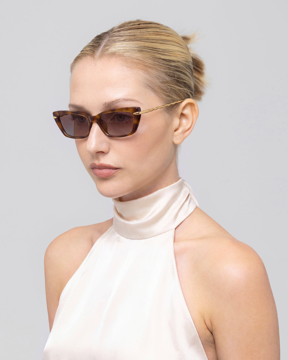 A blonde woman with a neat bun wears Sol II sunglasses by For Art’s Sake® and a shiny, high-neck, sleeveless cream top, posing against a plain light gray background.