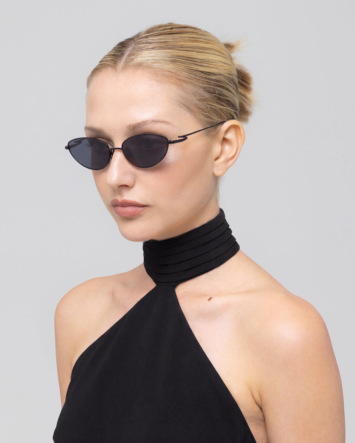 A woman with blonde hair in a bun wears For Art’s Sake® Ozanne I black sculptural sunglasses and a black high-neck halter top, facing slightly left against a plain light gray background.