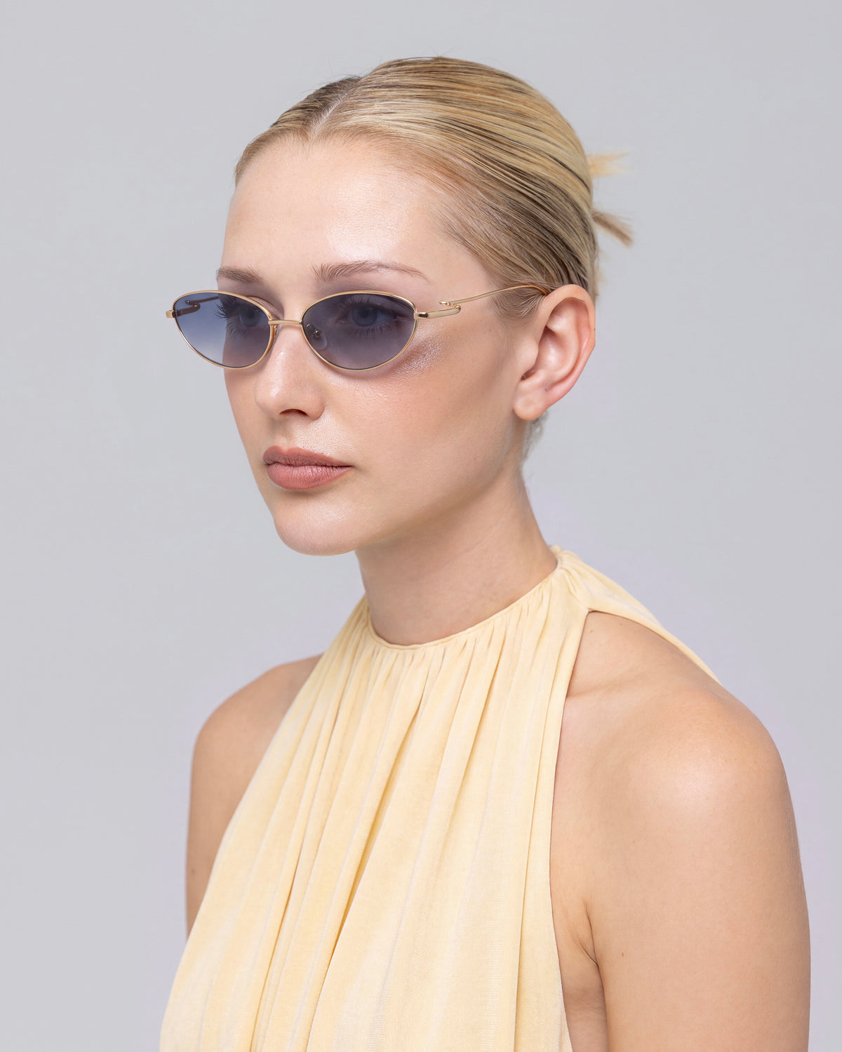 A woman with blonde hair in a low bun wears For Art’s Sake® Ozanne I small, jewellery-inspired blue sunglasses and a sleeveless light yellow halter dress, posing against a plain light gray background.