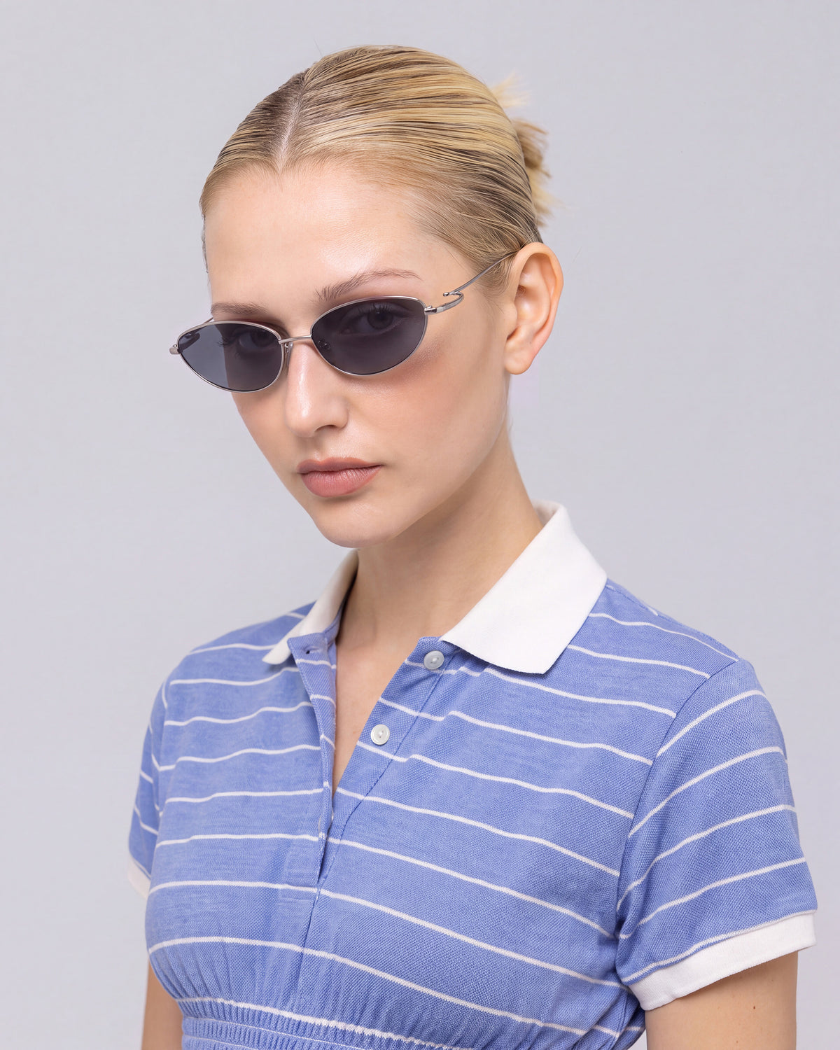 A woman with blonde hair in a bun wears For Art’s Sake® Ozanne I jewellery-inspired eyewear and a blue, short-sleeve, collared shirt with white stripes against a plain light gray background.