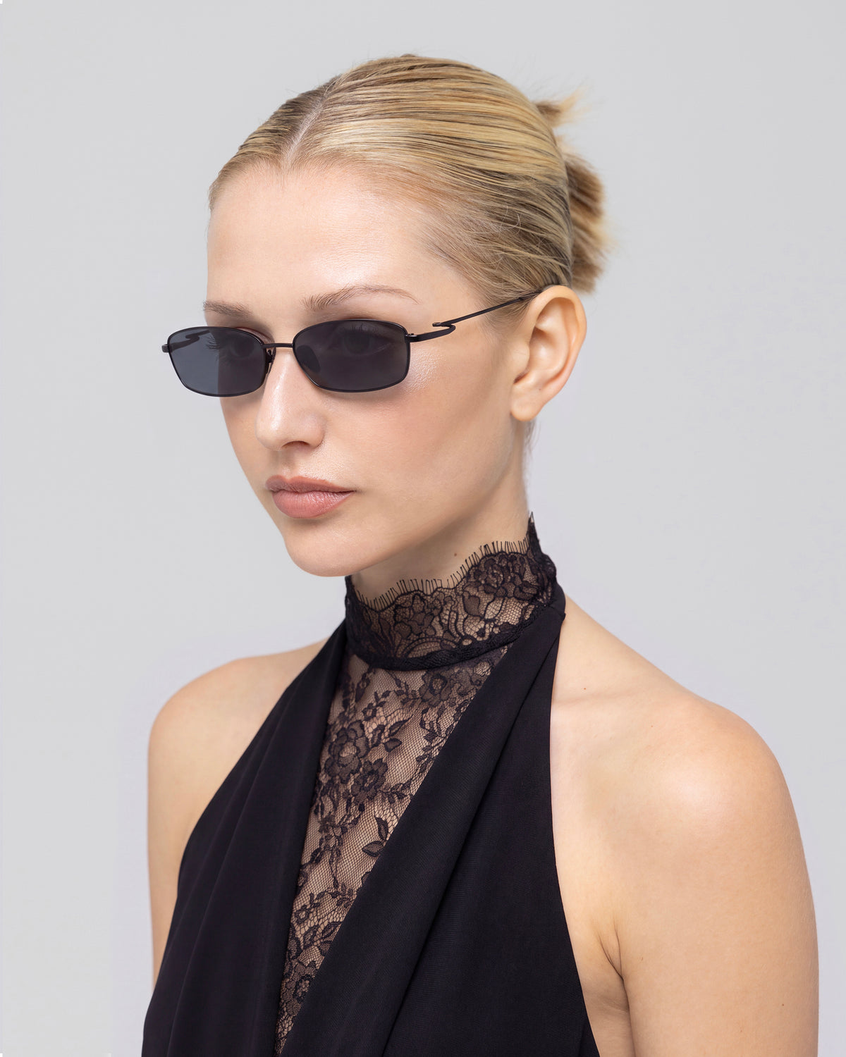 A woman with blond hair in a bun wears Ozanne II sunglasses by For Art’s Sake®, featuring sculptural looped temples, a sleeveless black outfit, and a lace high neck against a light gray background.