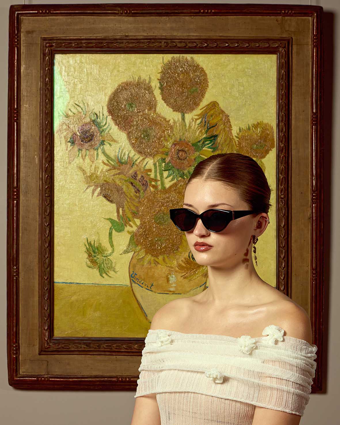 A woman in For Art's Sake® Vincent Van Gogh limited edition cat-eye sunglasses and an off-shoulder white dress poses by a framed sunflower painting.