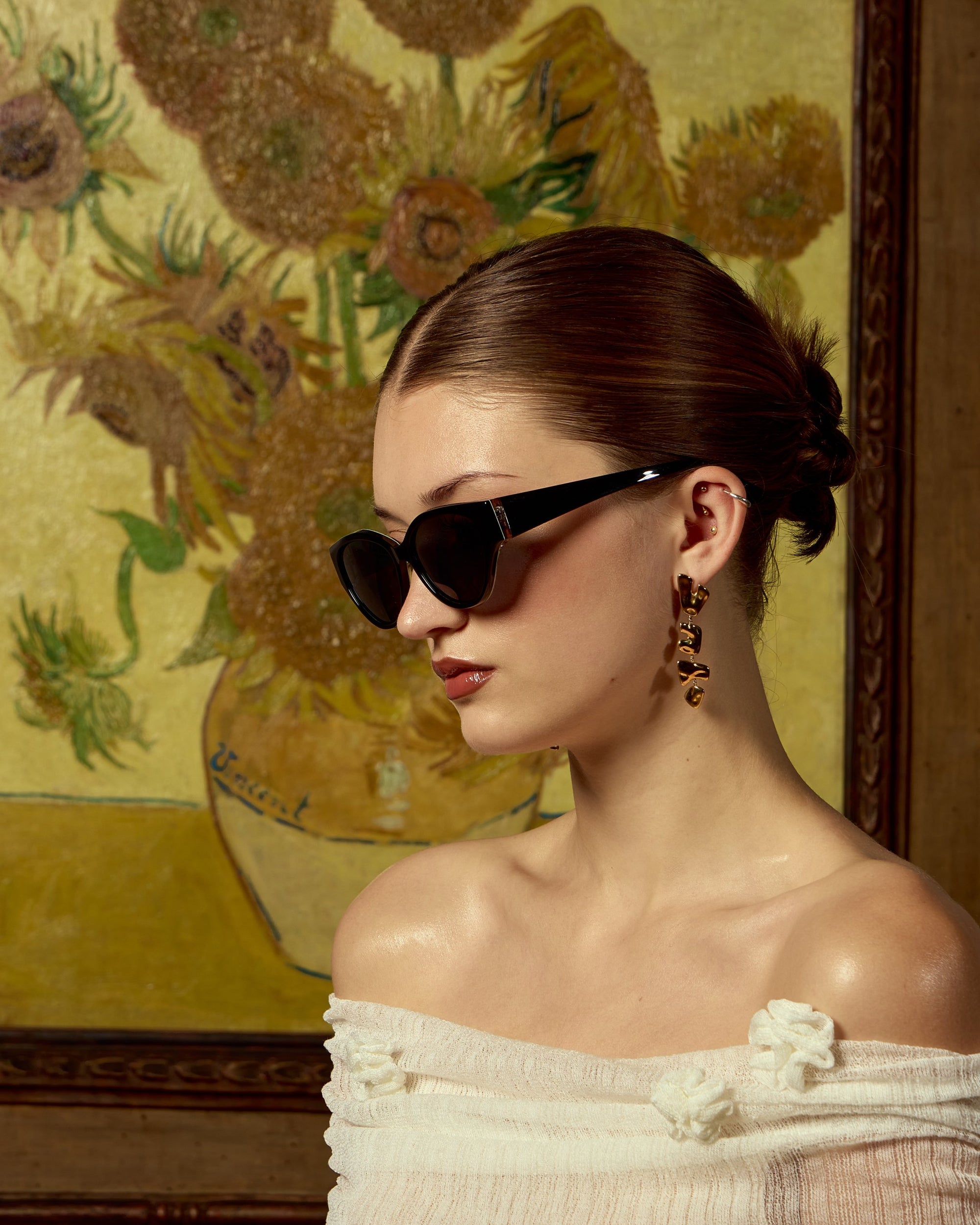A woman with slicked-back hair poses in profile, wearing For Art&#39;s Sake® Vincent Van Gogh limited edition cat-eye sunglasses, drop earrings, and an off-the-shoulder white top in front of a sunflower vase painting.
