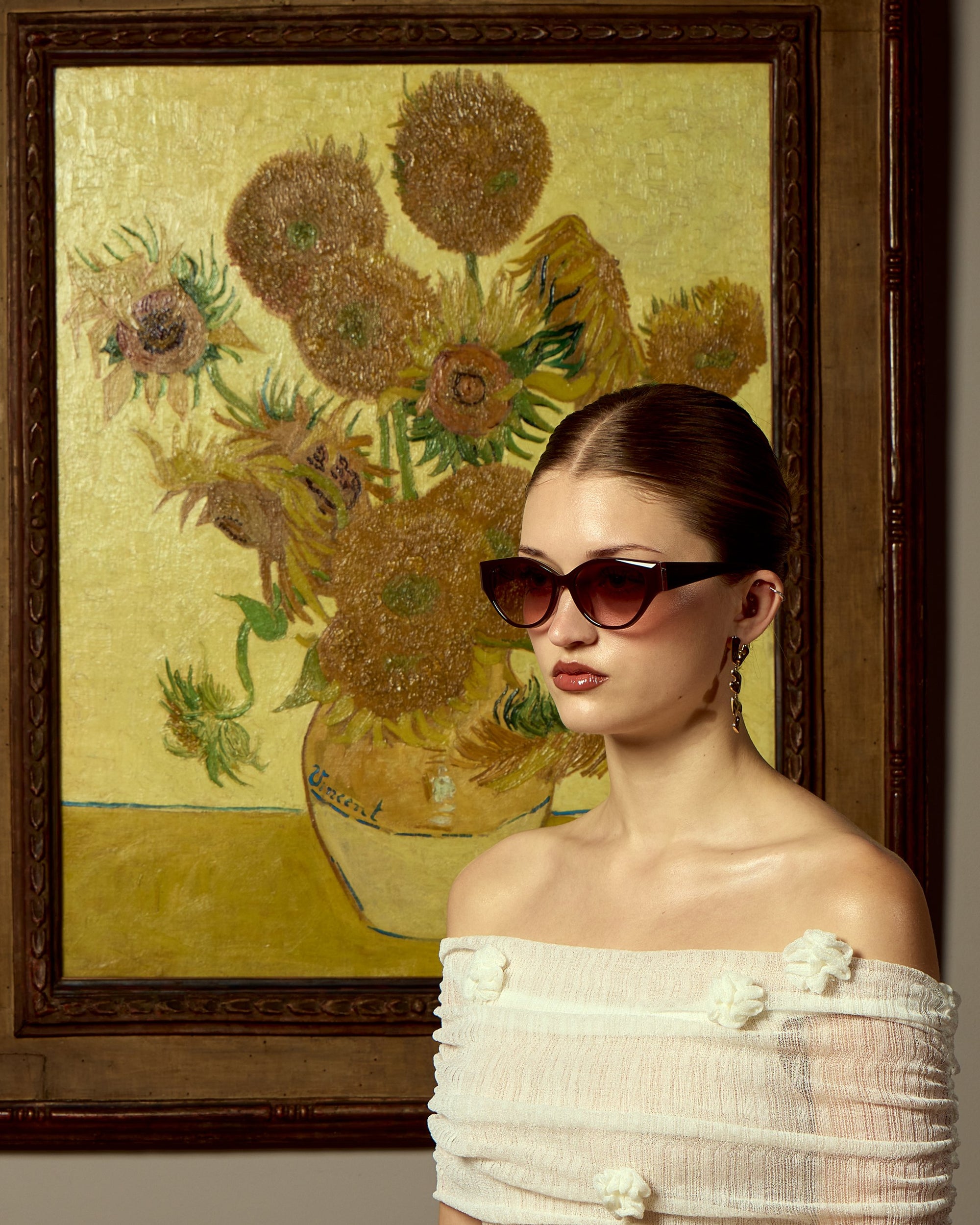 A woman wearing For Art's Sake® Vincent Van Gogh limited edition cat-eye sunglasses and an off-the-shoulder white top poses before a sunflower painting with a yellow background reminiscent of Van Gogh’s iconic style.