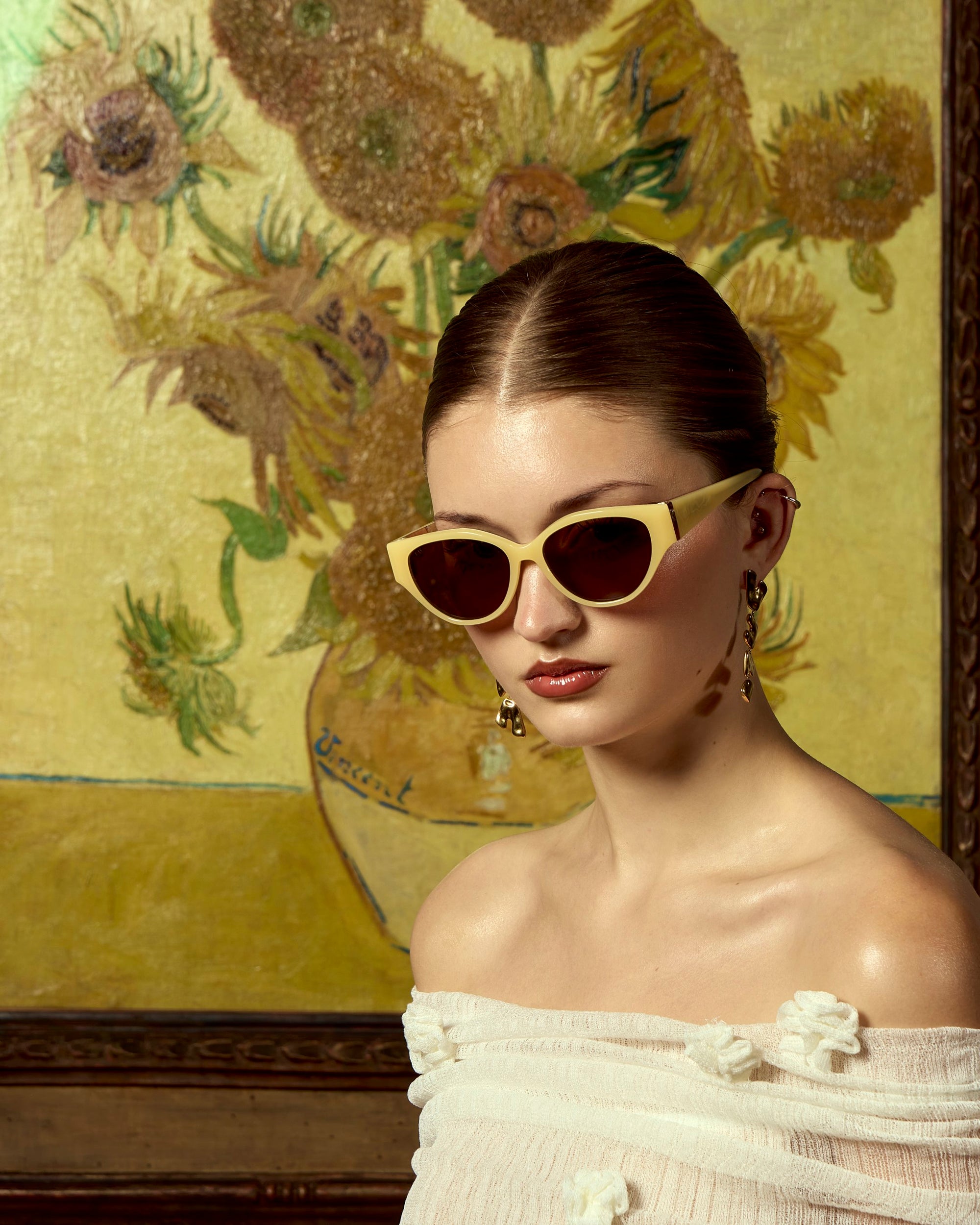 A woman in off-the-shoulder white attire and cream cat-eye sunglasses poses before a sunflower painting, her look completed with dangling earrings—showcasing the limited edition Vincent Van Gogh by For Art&#39;s Sake®.