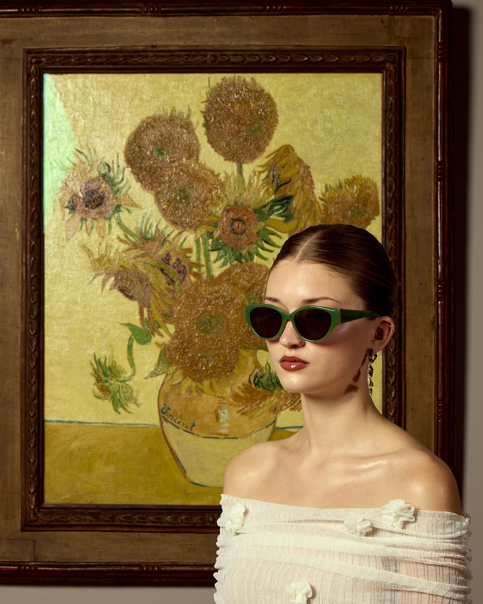 A woman wearing For Art's Sake® Vincent Van Gogh sunglasses and an off-the-shoulder white top stands before a framed sunflower painting with a vibrant yellow background, echoing the impressionist style.