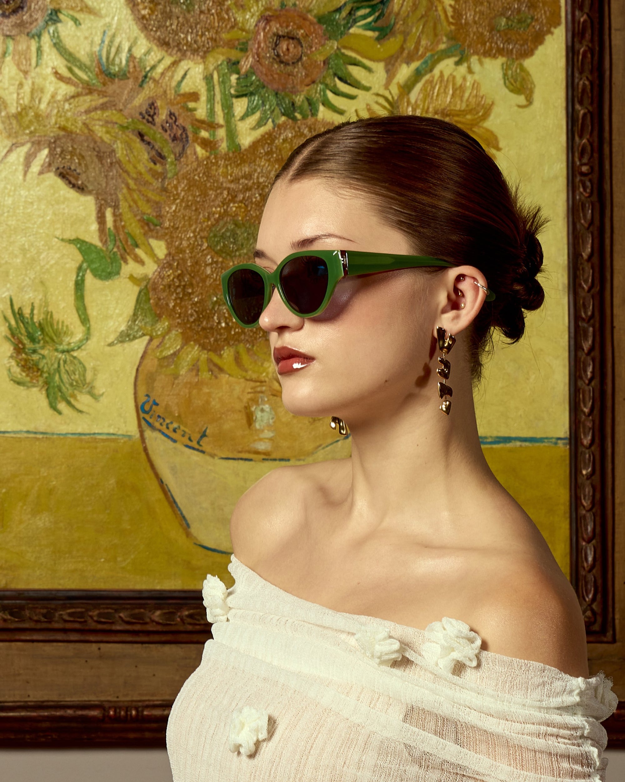 A woman in For Art&#39;s Sake® Vincent Van Gogh limited edition cat-eye sunglasses and a white off-shoulder top poses in profile before a framed sunflower painting, her hair tied back and wearing dangling earrings.