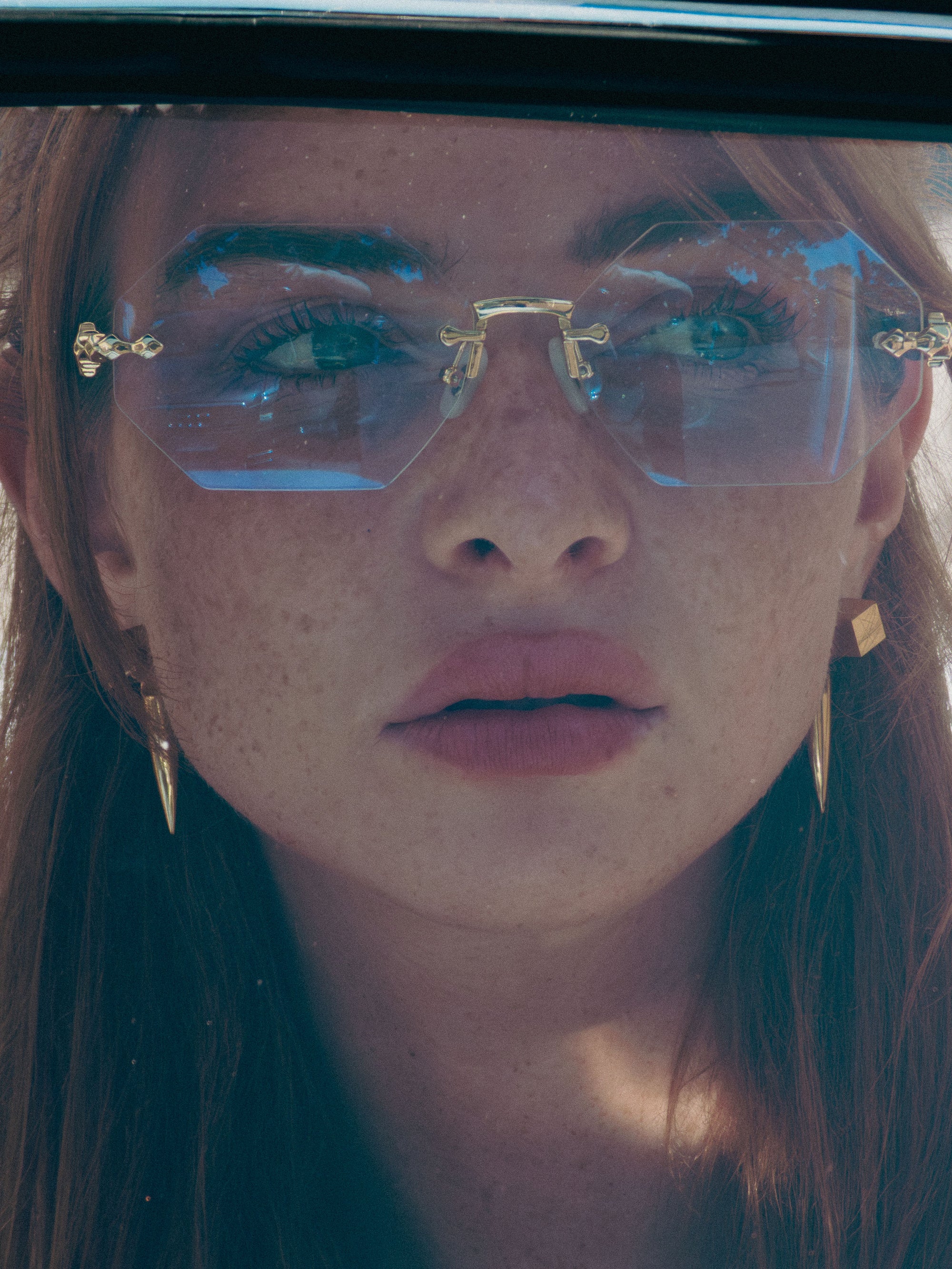 A young woman with freckles wears For Art's Sake® Hampton geometric sunglasses with faceted lenses and gold earrings, gazing upward as sunlight casts delicate shadows on her face.