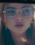 A young woman with freckles wears For Art's Sake® Hampton geometric sunglasses with faceted lenses and gold earrings, gazing upward as sunlight casts delicate shadows on her face.