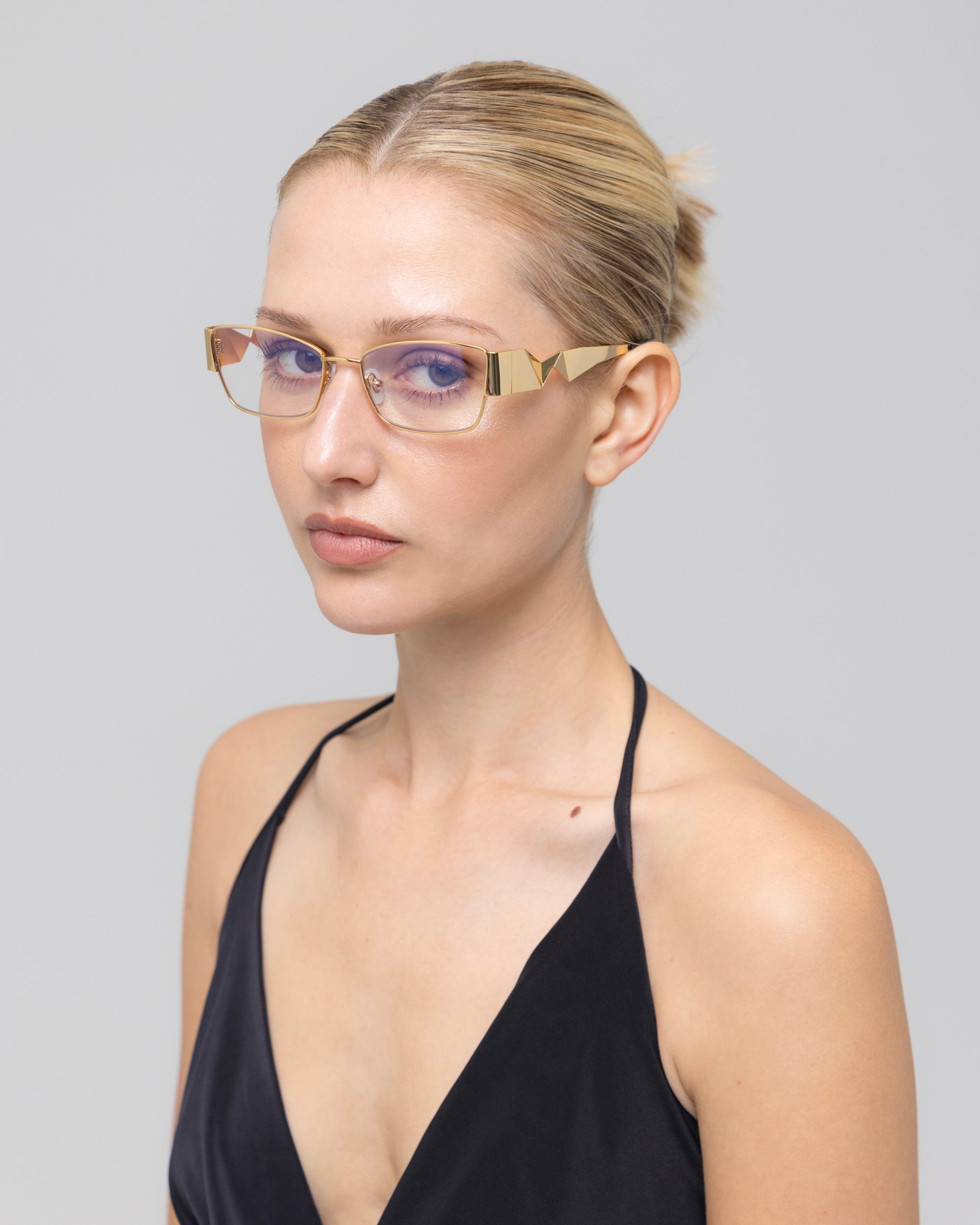 A woman with blonde hair in a sleek bun wears For Art's Sake® Origami geometric gold-rimmed glasses with a soft-square silhouette and a black halter top, looking at the camera against a plain light gray background.