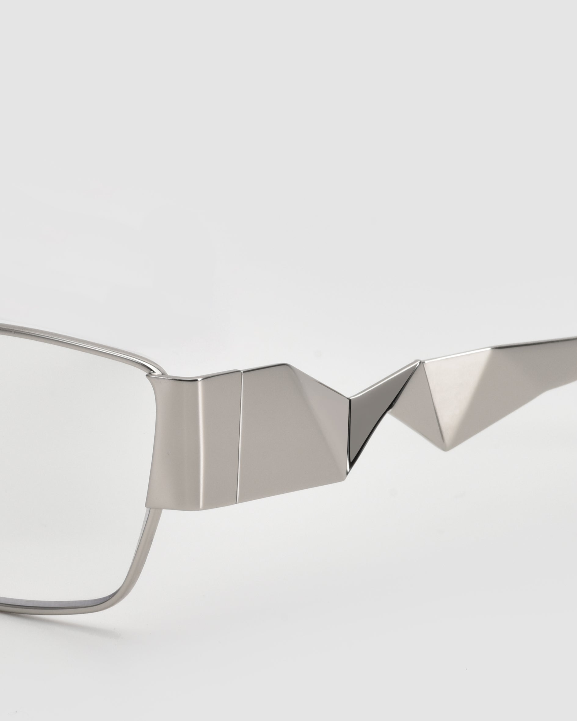 Close-up of For Art&#39;s Sake® Origami eyeglasses with pleated temples and an angular geometric metallic frame, set against a plain light gray background.