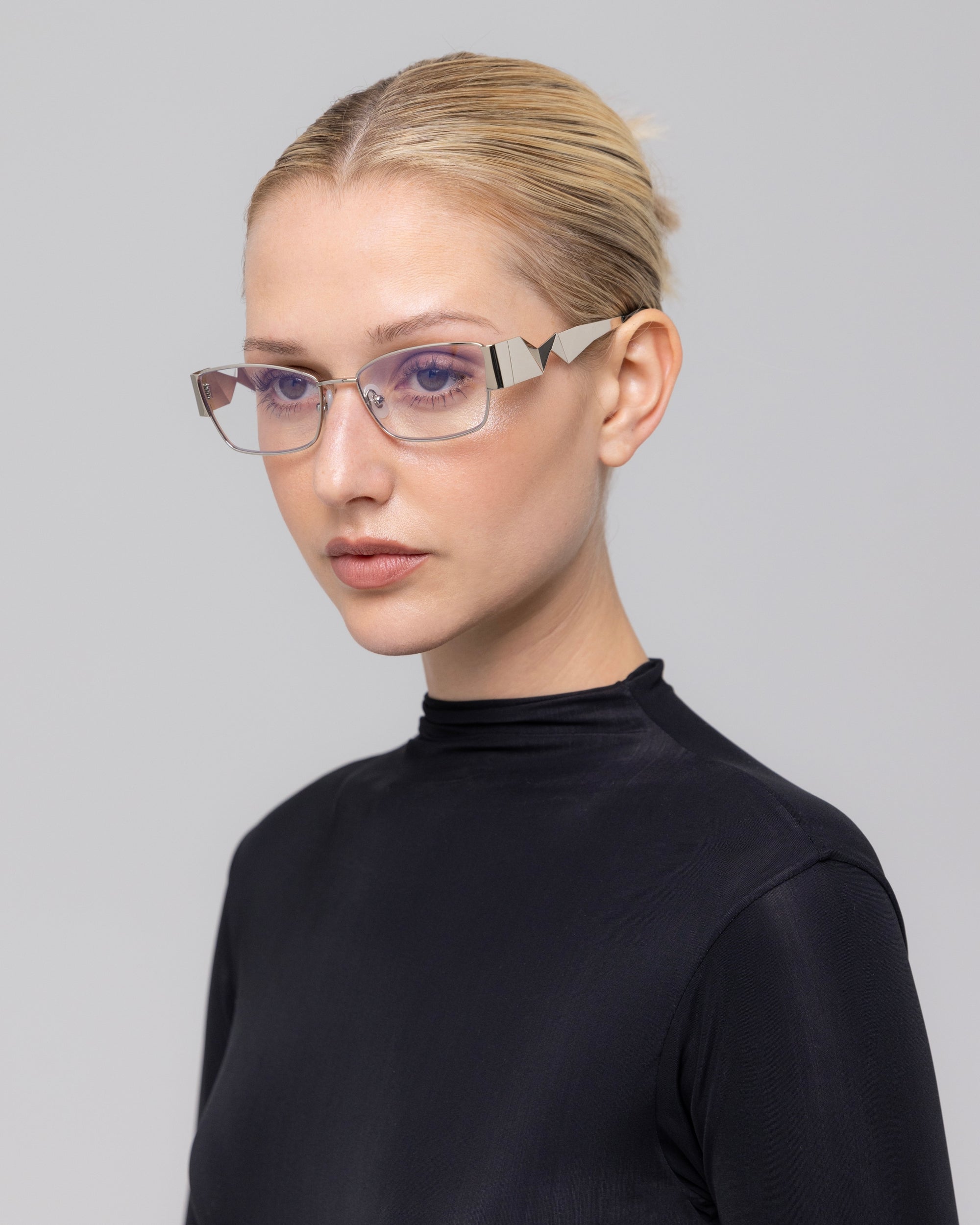 A blonde woman with her hair tied back wears For Art's Sake® Origami geometric eyeglasses and a fitted black turtleneck, posing against a plain light gray background.