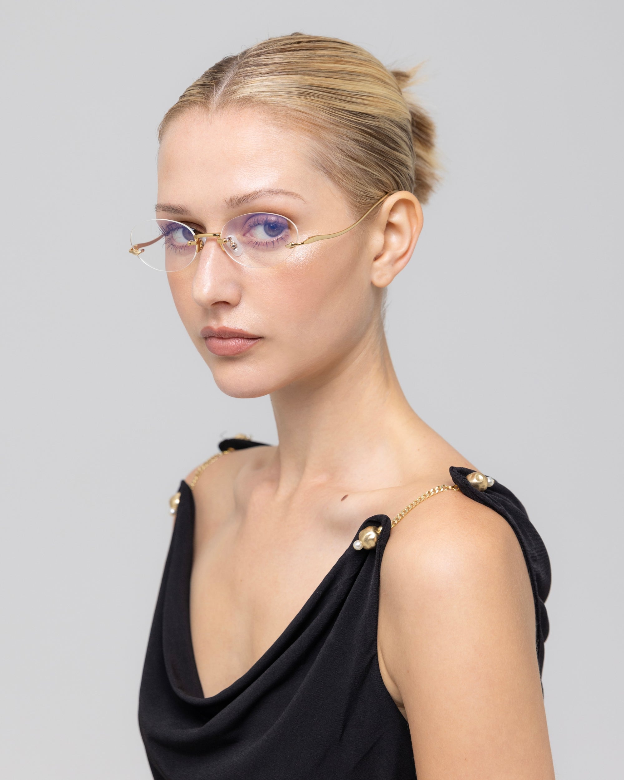A woman with blonde hair in a neat bun wears For Art's Sake® Waver minimalistic eyeglass frames and a sleeveless black top with gold strap details, gazing slightly to the side against a plain light gray background.