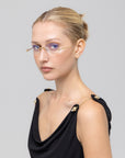 A woman with blonde hair in a neat bun wears For Art's Sake® Waver minimalistic eyeglass frames and a sleeveless black top with gold strap details, gazing slightly to the side against a plain light gray background.
