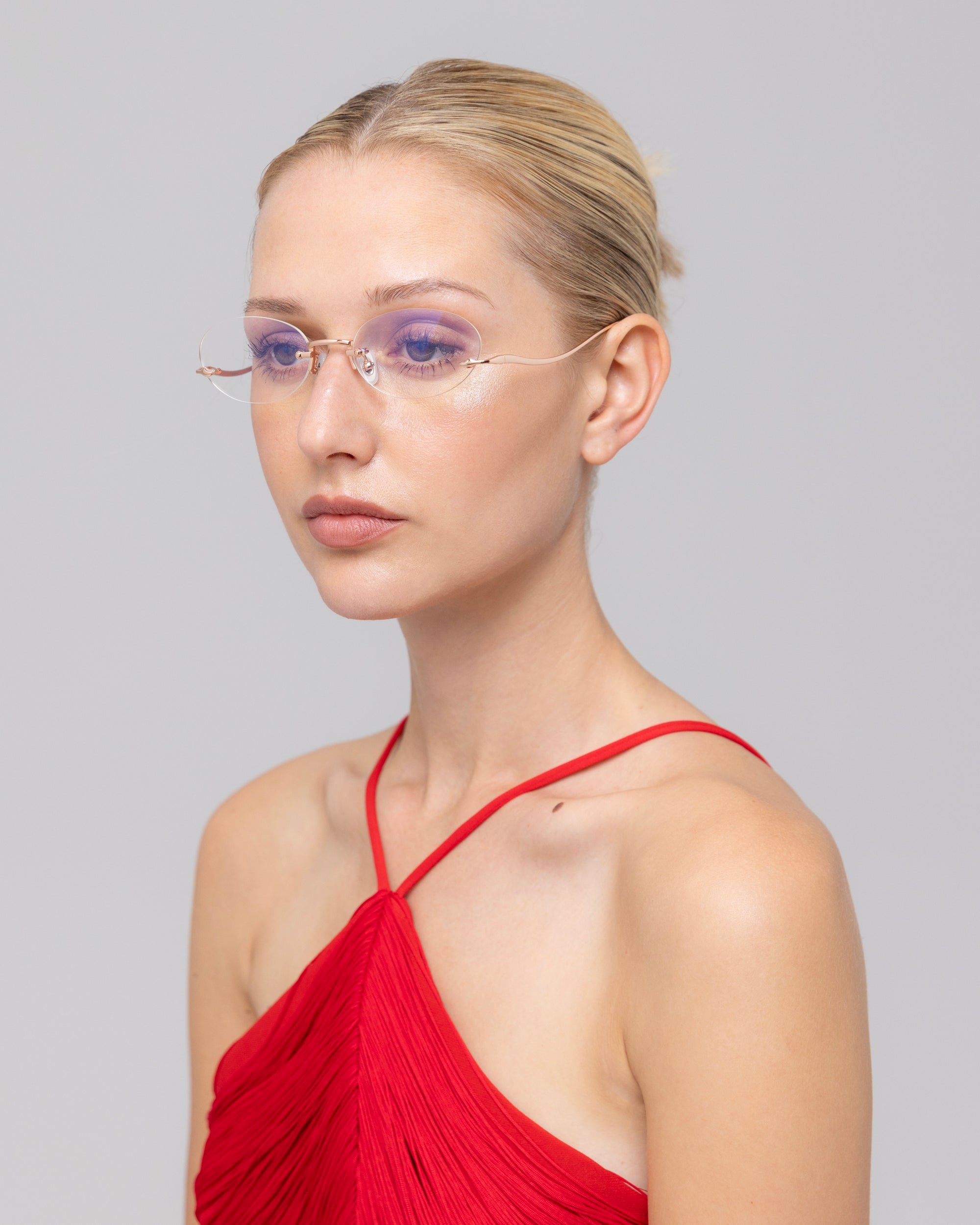 A woman with blonde hair in a bun wears For Art's Sake® Waver rimless glasses and a red pleated halter-neck top. She poses with a neutral expression against a plain light gray background.