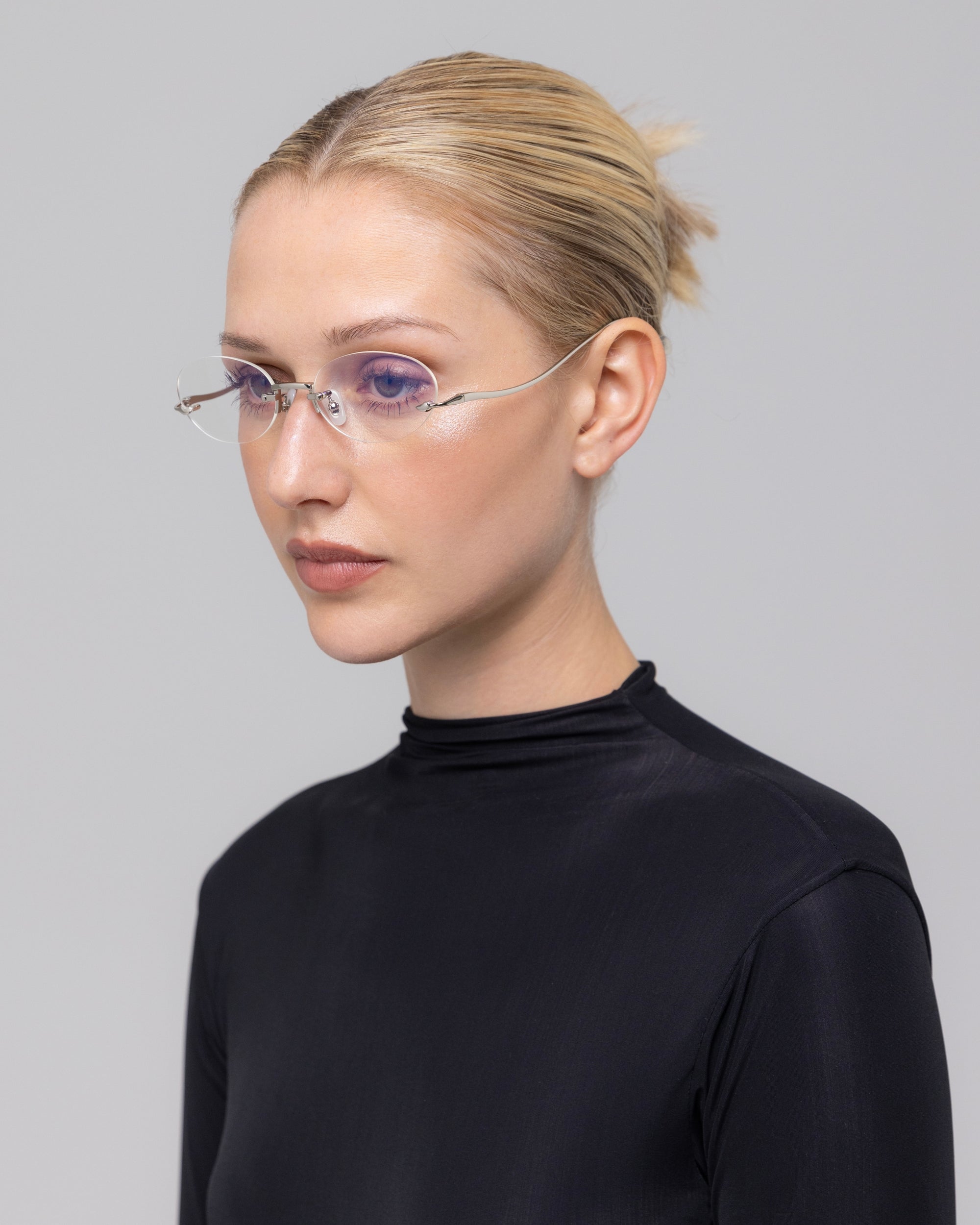 A woman with blonde hair in a neat bun models For Art's Sake® Waver minimalistic eyeglass frames and a black high-neck top, looking slightly to the side against a plain light gray background.