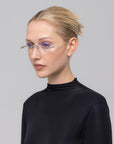 A woman with blonde hair in a neat bun models For Art's Sake® Waver minimalistic eyeglass frames and a black high-neck top, looking slightly to the side against a plain light gray background.