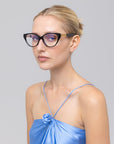 A woman with a sleek blonde bun wears The Act black cat-eye glasses by For Art's Sake® and a shiny light blue spaghetti strap top, looking slightly to the side against a plain gray background.