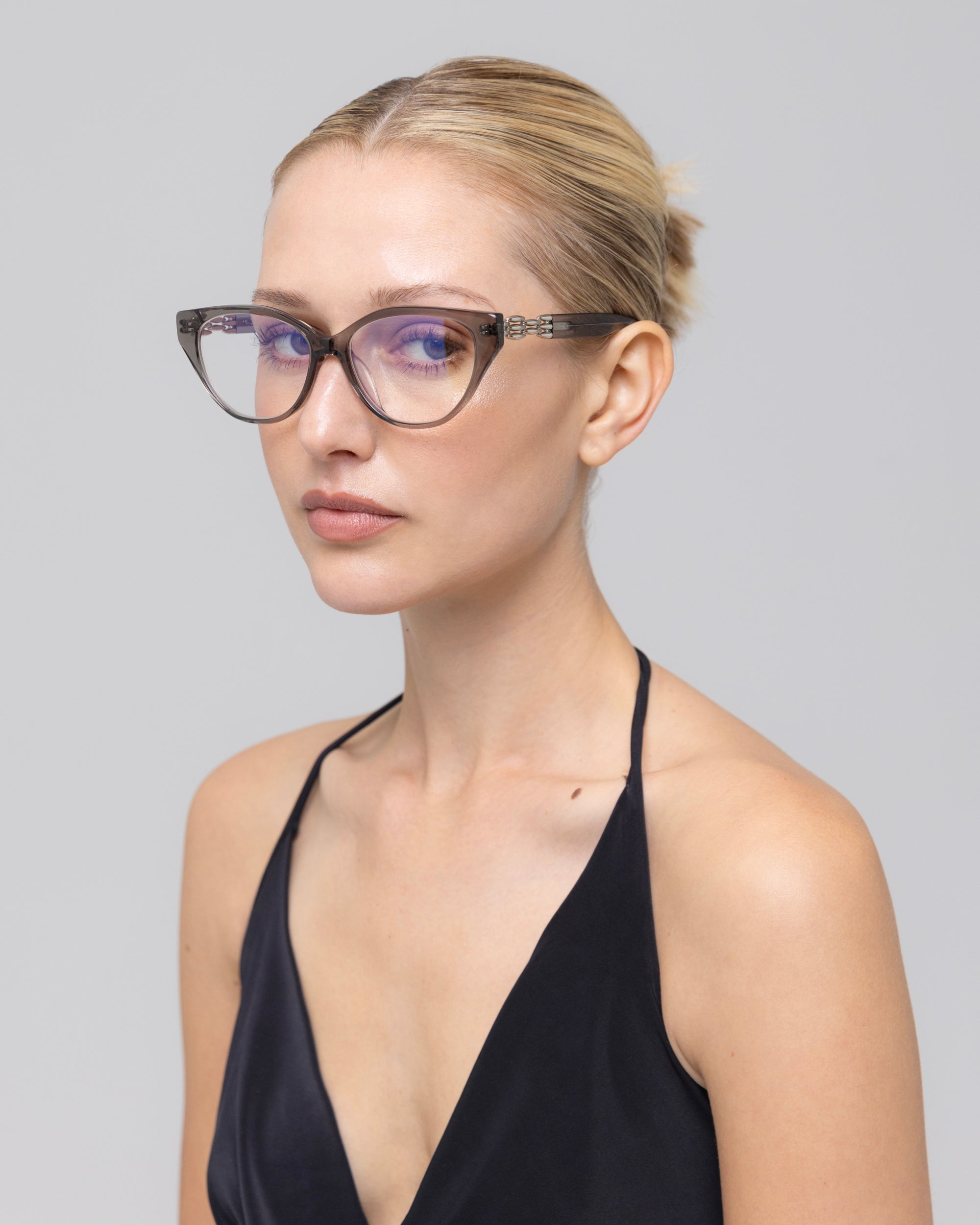 A woman with blonde hair tied back wears The Act eyeglasses by For Art's Sake®, featuring a lightweight clear frame and a sleeveless black top, looking calmly at the camera against a plain light gray background.
