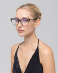 A woman with blonde hair tied back wears The Act eyeglasses by For Art's Sake®, featuring a lightweight clear frame and a sleeveless black top, looking calmly at the camera against a plain light gray background.