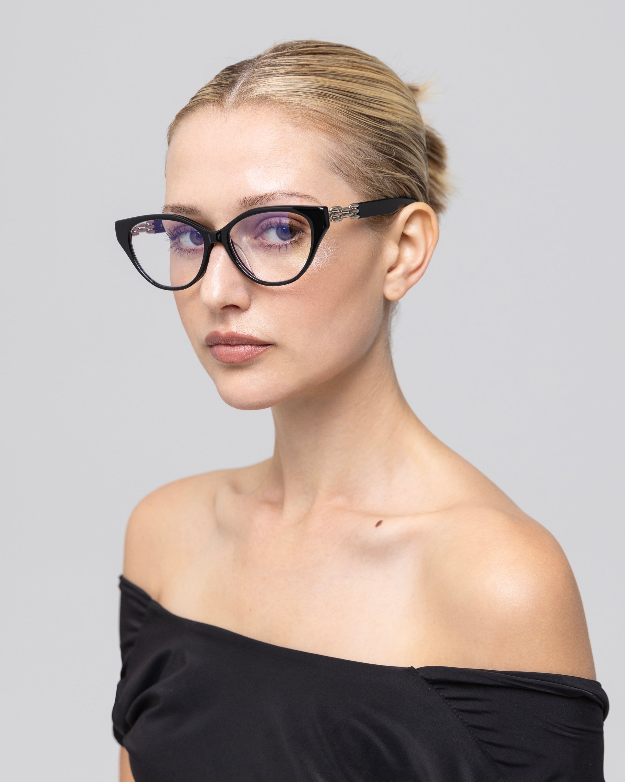 A woman with blonde hair in a neat bun models The Act minimalist eyeglass frames by For Art's Sake®, paired with an off-the-shoulder black top, gazing sideways against a plain light gray background.