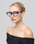 A woman with blonde hair in a neat bun models The Act minimalist eyeglass frames by For Art's Sake®, paired with an off-the-shoulder black top, gazing sideways against a plain light gray background.