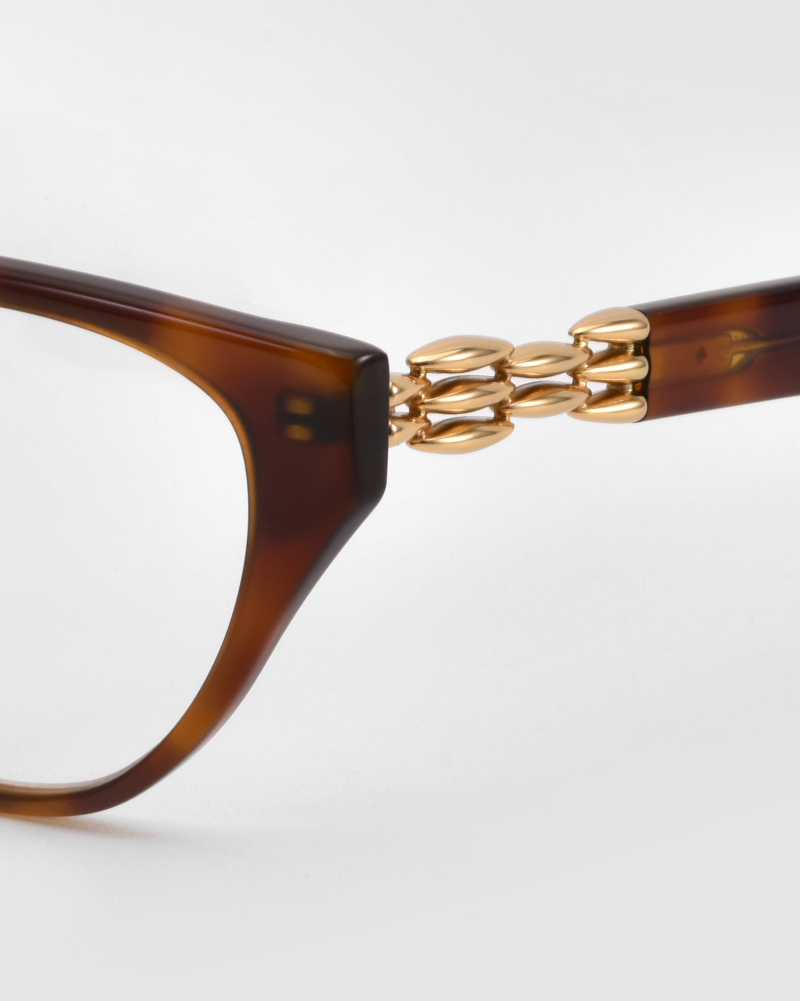 Close-up of &quot;The Act&quot; brown cat-eye sunglasses by For Art&#39;s Sake®, showcasing a gold chain link detail at the hinge with a 3D weaved metal look, set against a white background.