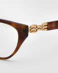 Close-up of "The Act" brown cat-eye sunglasses by For Art's Sake®, showcasing a gold chain link detail at the hinge with a 3D weaved metal look, set against a white background.