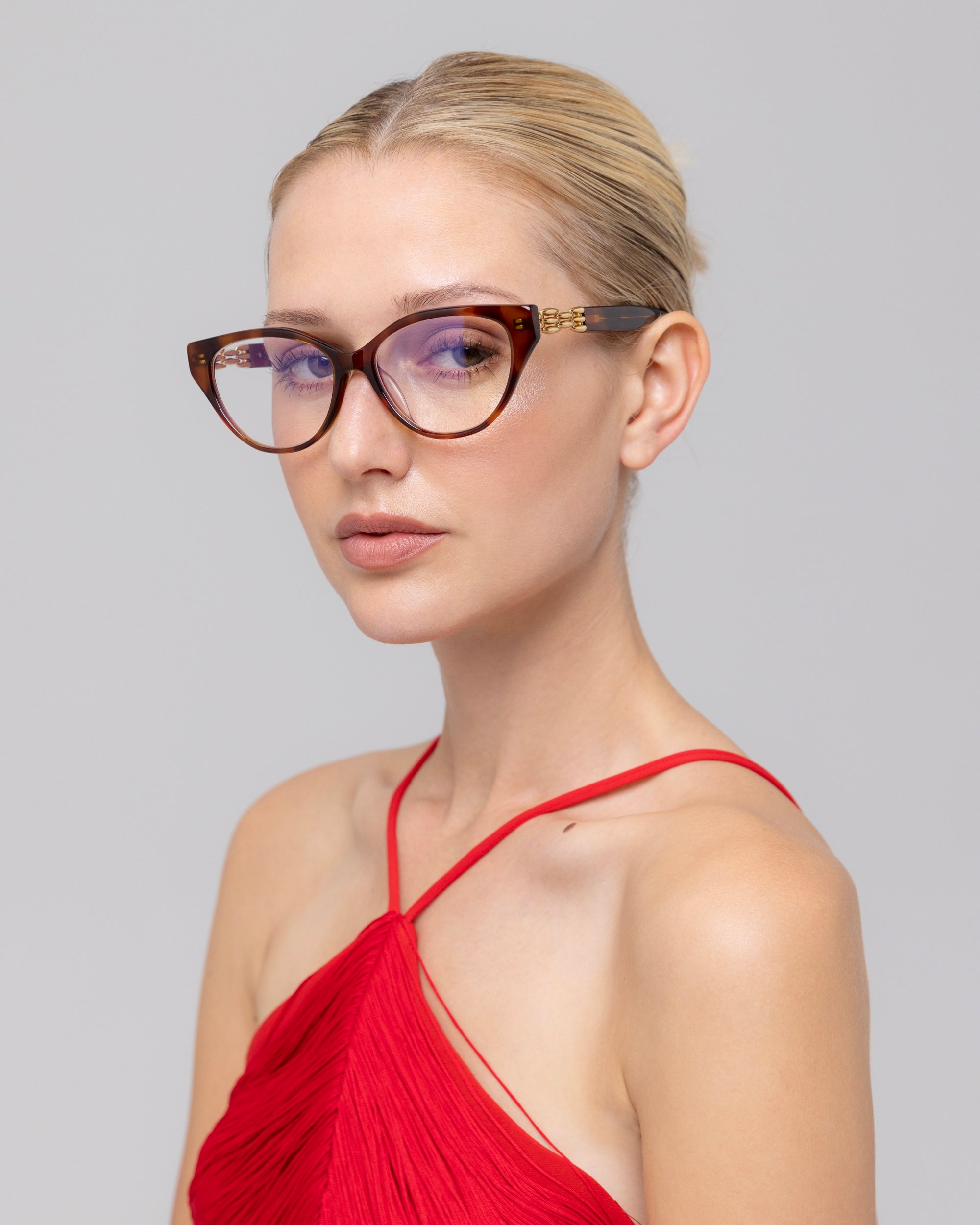 A woman with blonde hair in a sleek bun models The Act cat-eye eyewear by For Art's Sake®, paired with a red halter-neck dress against a plain light gray background.