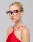 A woman with blonde hair in a sleek bun models The Act cat-eye eyewear by For Art's Sake®, paired with a red halter-neck dress against a plain light gray background.