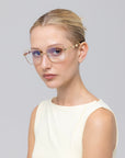 A woman with blonde hair in a neat bun wears For Art's Sake® Fluid eyeglasses—large, metal frames in a soft-square shape—and a sleeveless off-white top, gazing sideways against a plain light grey background.