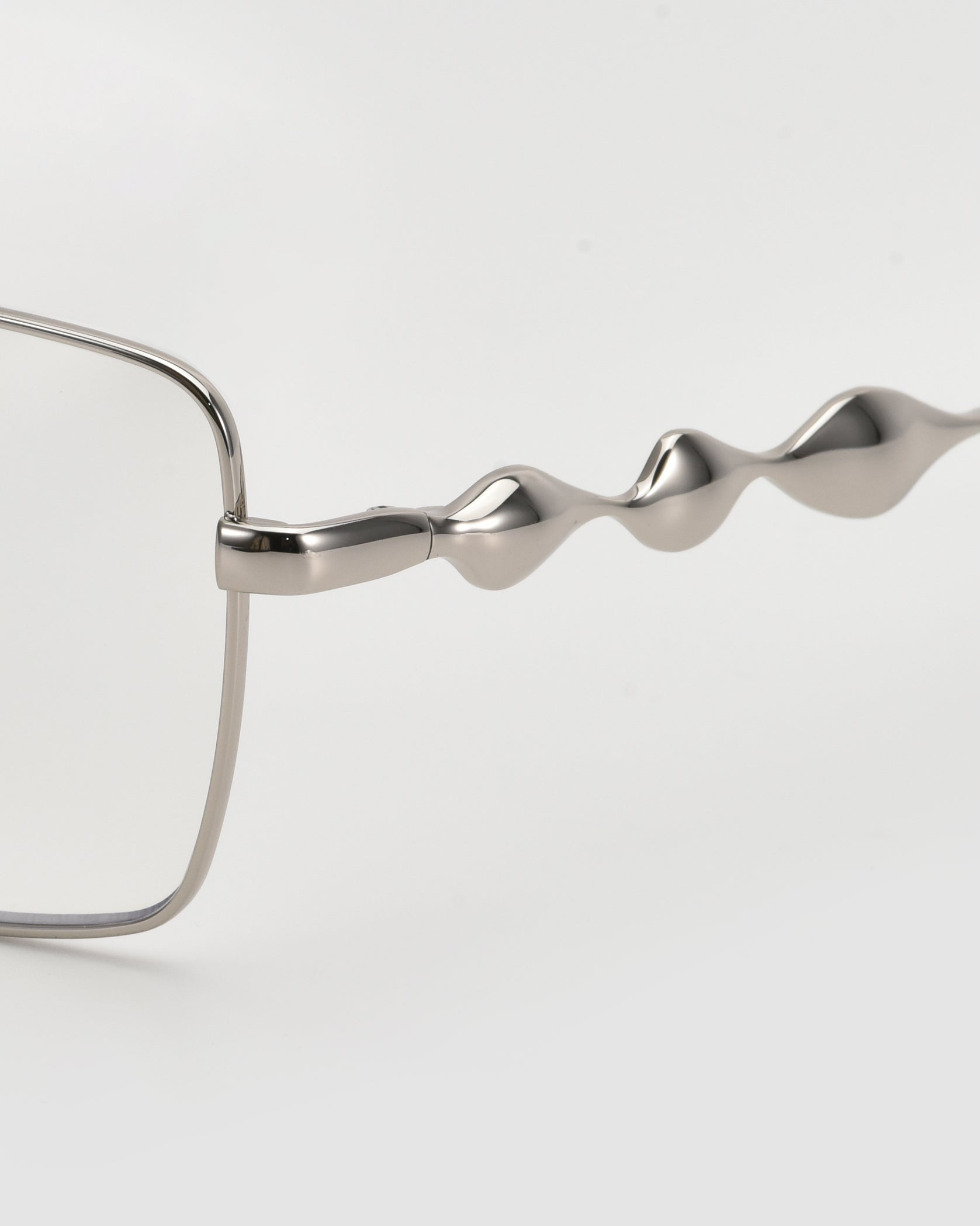 Close-up of For Art&#39;s Sake® Fluid: a silver eyeglass frame with a 3D fluid design on the temple arm and soft-square lenses, shown against a plain white background.