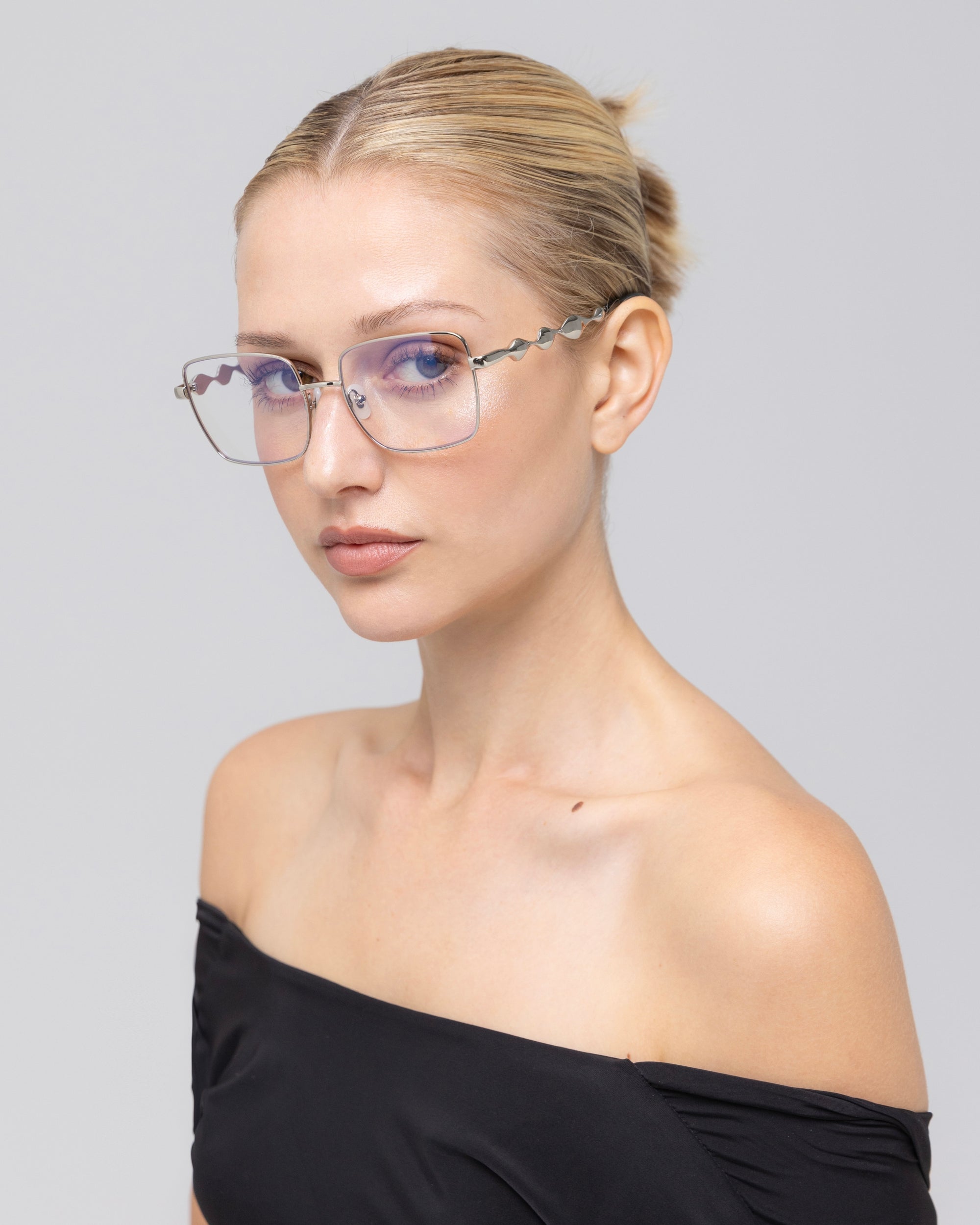 A woman with blonde hair in a neat bun wears Fluid rimless eyeglasses by For Art's Sake® and an off-the-shoulder black top, looking at the camera with a neutral expression against a plain light gray background.