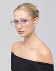 A woman with blonde hair in a neat bun wears Fluid rimless eyeglasses by For Art's Sake® and an off-the-shoulder black top, looking at the camera with a neutral expression against a plain light gray background.