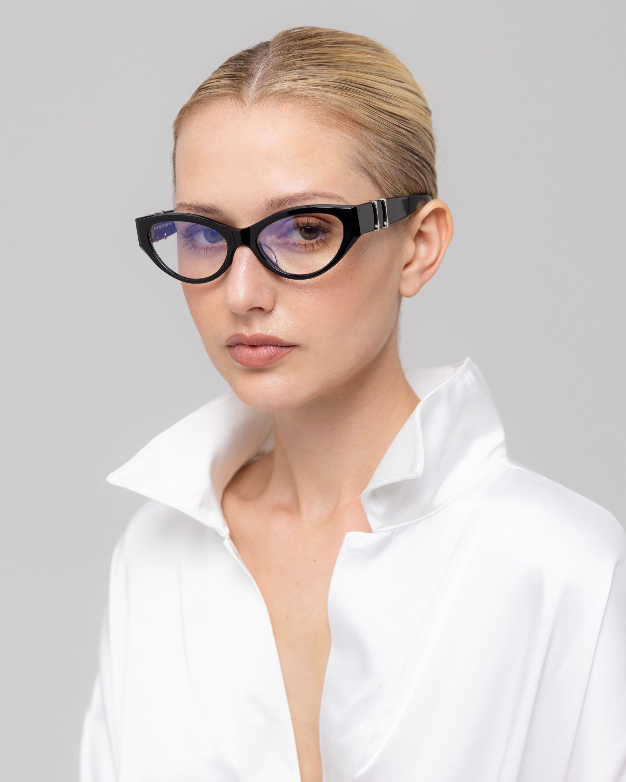 A woman with blonde hair in a sleek bun wears For Art's Sake® Halo(X) black cat-eye sunglasses and a white satin blouse with an open collar, posing against a plain light gray background.