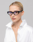A woman with blonde hair in a sleek bun wears For Art's Sake® Halo(X) black cat-eye sunglasses and a white satin blouse with an open collar, posing against a plain light gray background.