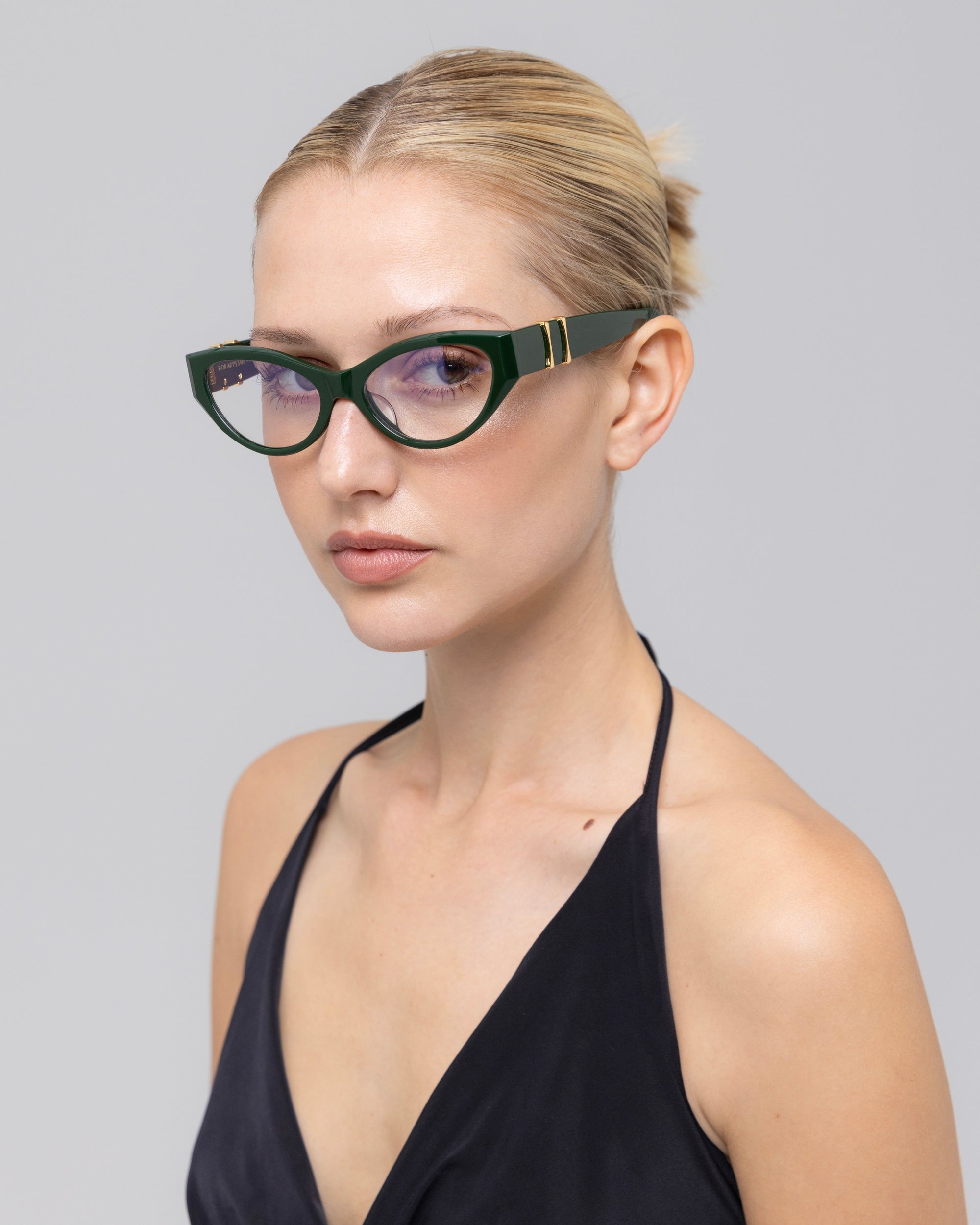 A woman with blonde hair in a bun wears For Art's Sake® Halo(X) bold cat-eye glasses and a black halter top, confidently showcasing chic everyday eyewear against a plain light gray background.