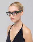 A woman with blonde hair in a bun wears For Art's Sake® Halo(X) bold cat-eye glasses and a black halter top, confidently showcasing chic everyday eyewear against a plain light gray background.