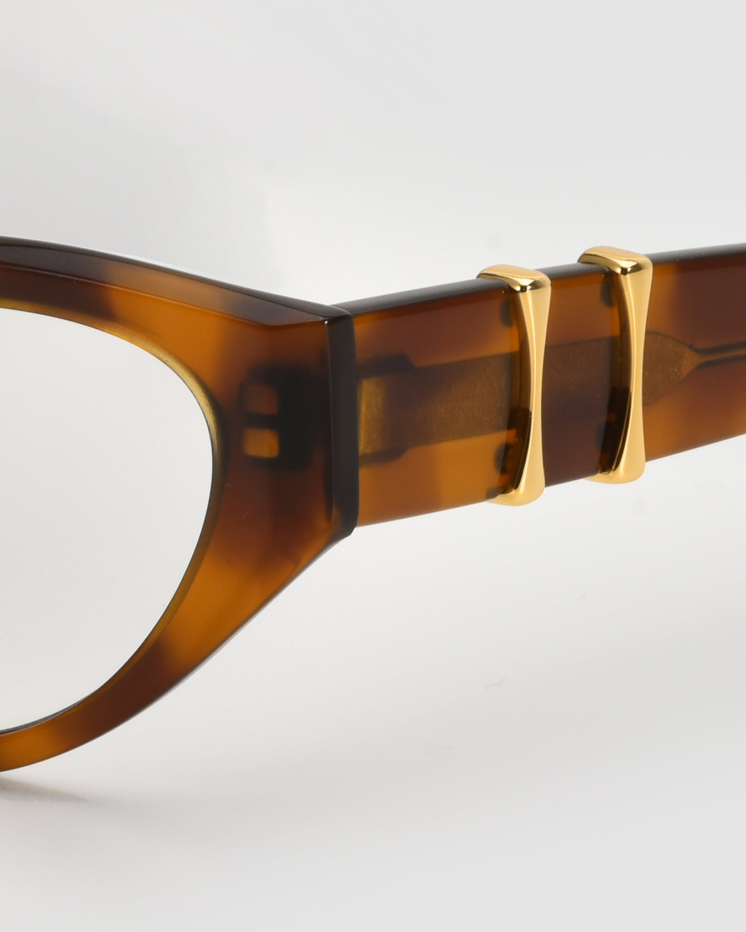 Close-up of the side of the Halo(X) sculptural brown tortoiseshell acetate frame by For Art&#39;s Sake®, featuring two gold metal accents on the temple against a light gray background.