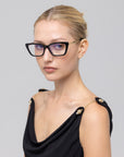 A woman with blonde hair tied back wears For Art's Sake® Harmony(X) black cat-eye sunglasses and a sleeveless black dress with gold buttons, looking at the camera against a light gray background.