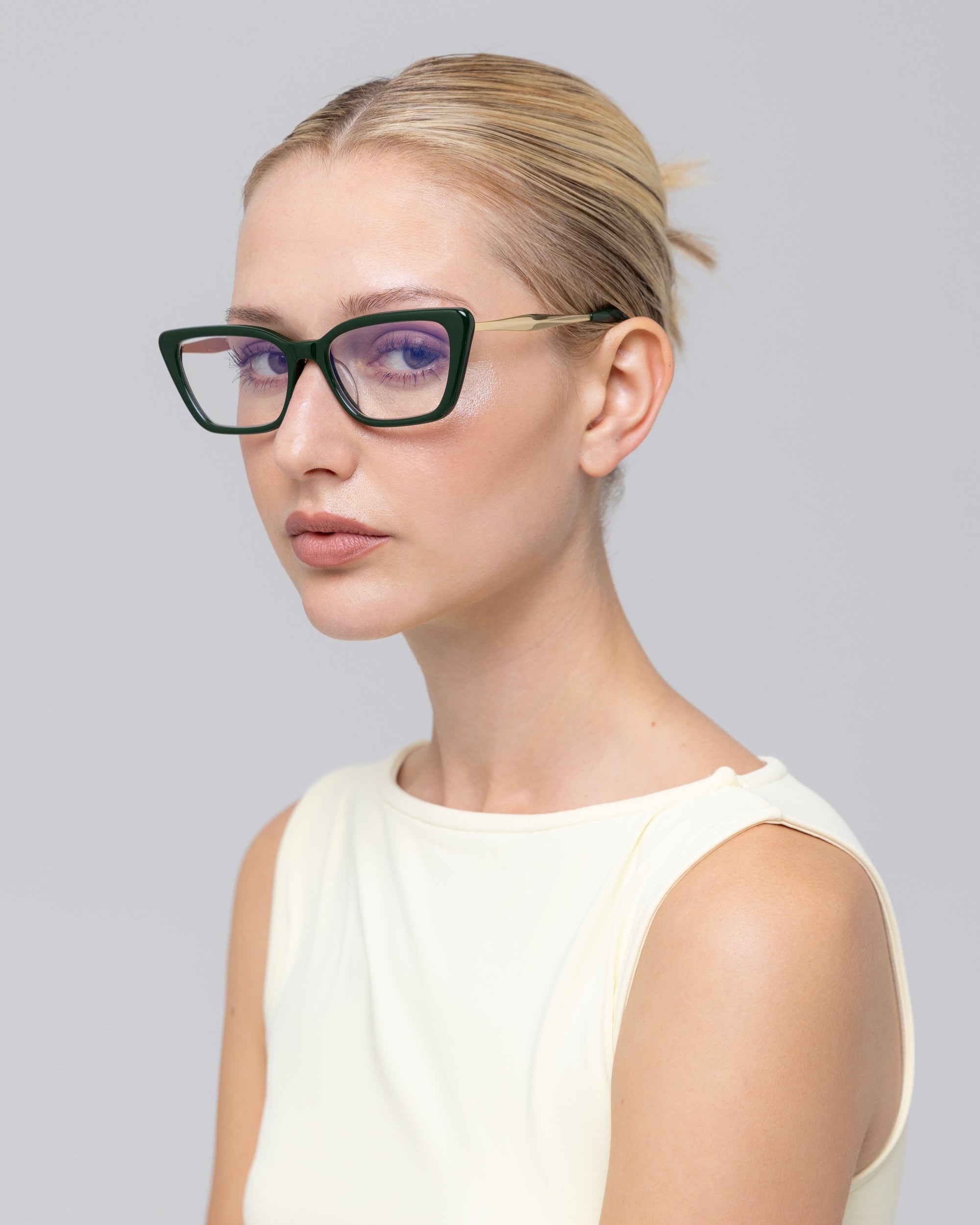 A woman with blonde hair pulled back wears For Art's Sake® Harmony(X) black cat-eye glasses with sculptural temples and a sleeveless off-white top, looking to the side against a plain light gray background.