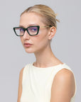 A woman with blonde hair pulled back wears For Art's Sake® Harmony(X) black cat-eye glasses with sculptural temples and a sleeveless off-white top, looking to the side against a plain light gray background.