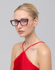 A woman with blonde hair tied back wears For Art's Sake® Harmony(X) brown square eyeglasses with sculptural temples and a red halter top, posing against a plain light gray background.