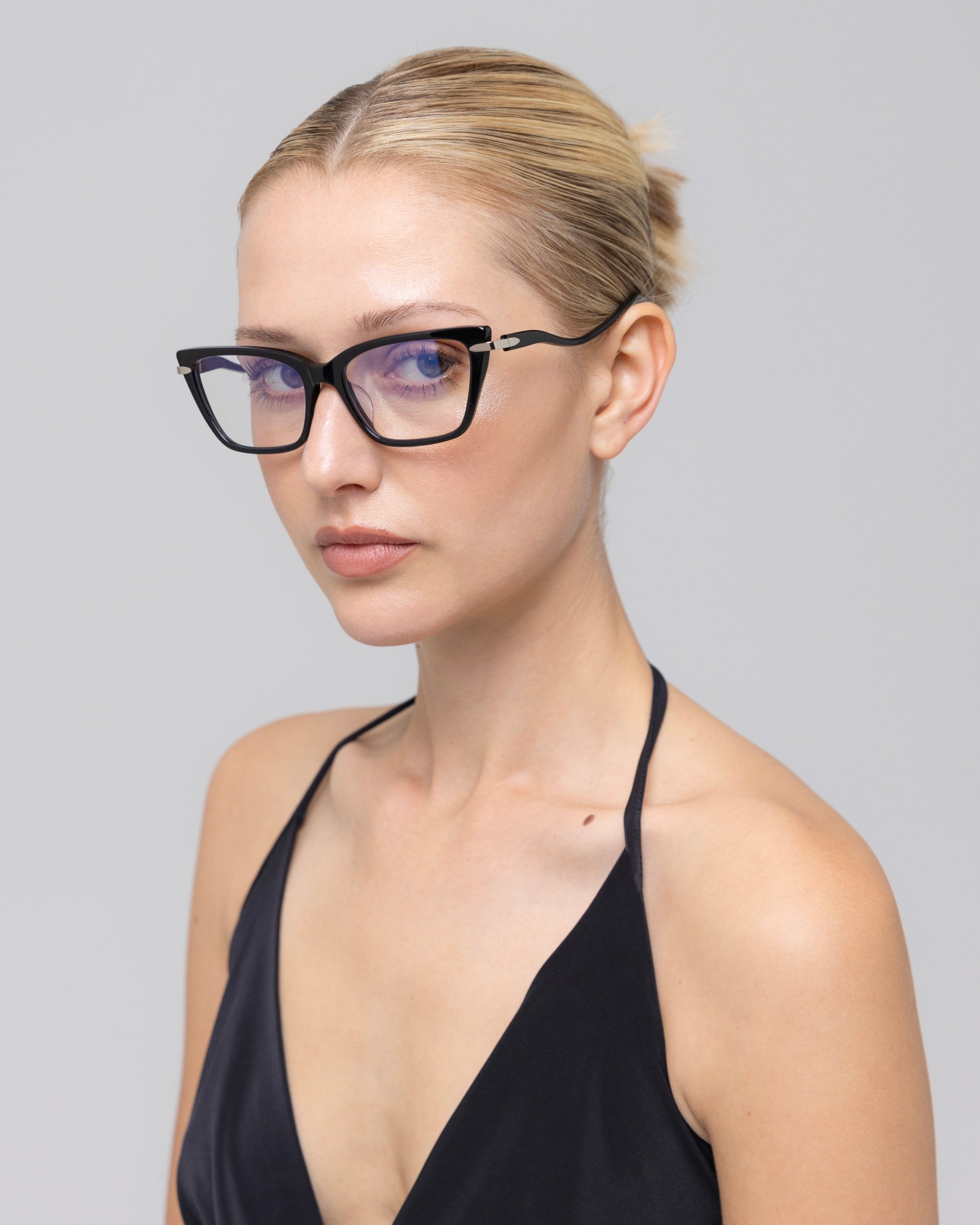 A woman with blonde hair in a neat bun wears For Art's Sake® Motion I black cat-eye glasses with acetate temples and a black halter top, gazing slightly to the side against a plain light gray background.