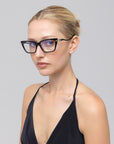 A woman with blonde hair in a neat bun wears For Art's Sake® Motion I black cat-eye glasses with acetate temples and a black halter top, gazing slightly to the side against a plain light gray background.